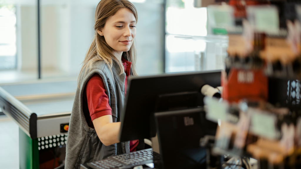 A professional cashier smiling at a checkout counter while using a Point of Sale (POS) system in a retail store.